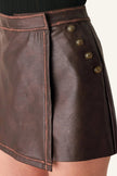 Close-up of a dark brown faux-leather wrap mini skirt with decorative gold-tone buttons on the side, shown on a model.