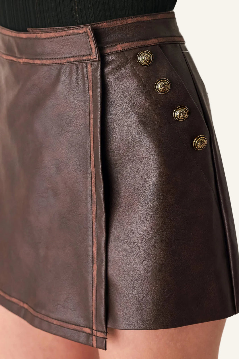 Close-up of a dark brown faux-leather wrap mini skirt with decorative gold-tone buttons on the side, shown on a model.