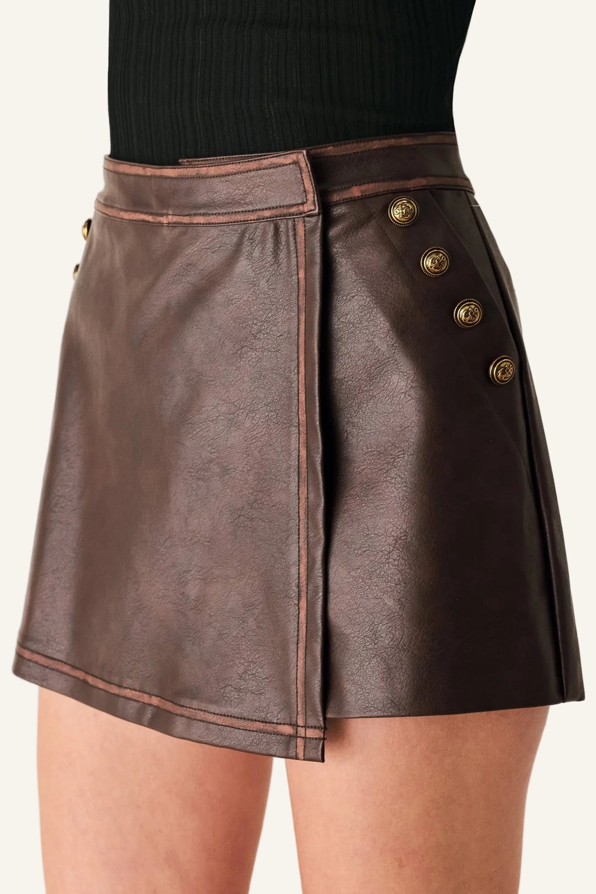 A close-up of a model wearing a dark brown faux-leather wrap mini skirt with gold-tone decorative buttons on the side, paired with a black ribbed top.