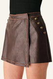 A close-up of a model wearing a dark brown faux-leather wrap mini skirt with gold-tone decorative buttons on the side, paired with a black ribbed top.
