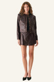 A model stands against a plain light gray studio background wearing a dark brown faux-leather jacket and matching mini skirt set with gold-tone hardware and black pointed-toe heels.