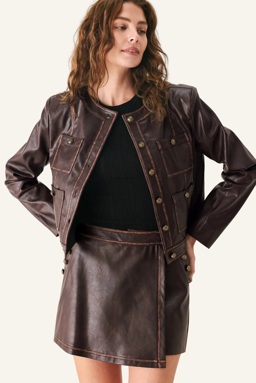 A woman wearing a dark brown faux leather jacket and matching mini skirt over a black ribbed top, photographed against a light gray studio background.