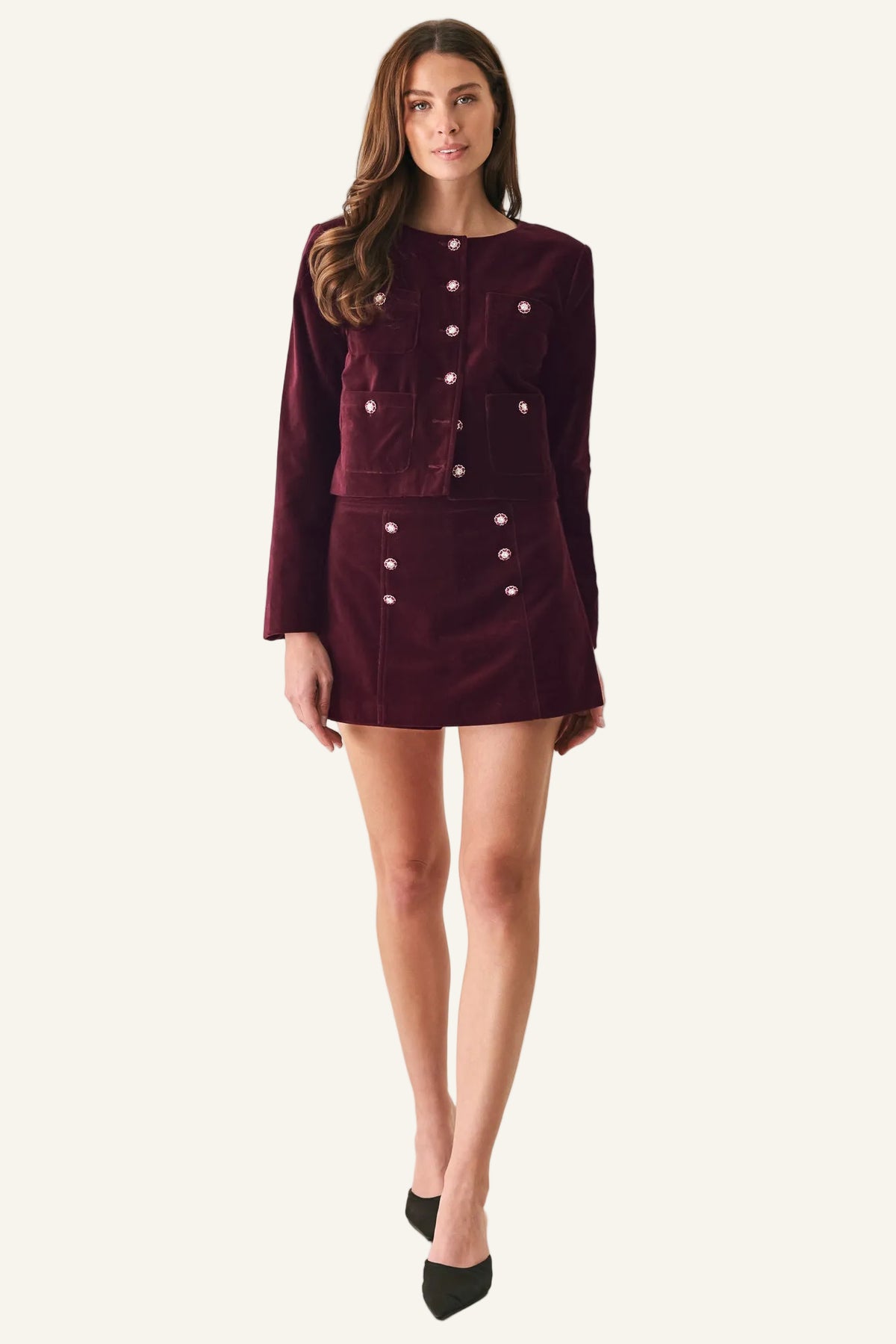 A woman modeling a matching burgundy velvet jacket and mini skirt set with silver-tone decorative buttons, paired with black pointed-toe heels, against a plain off-white studio background.