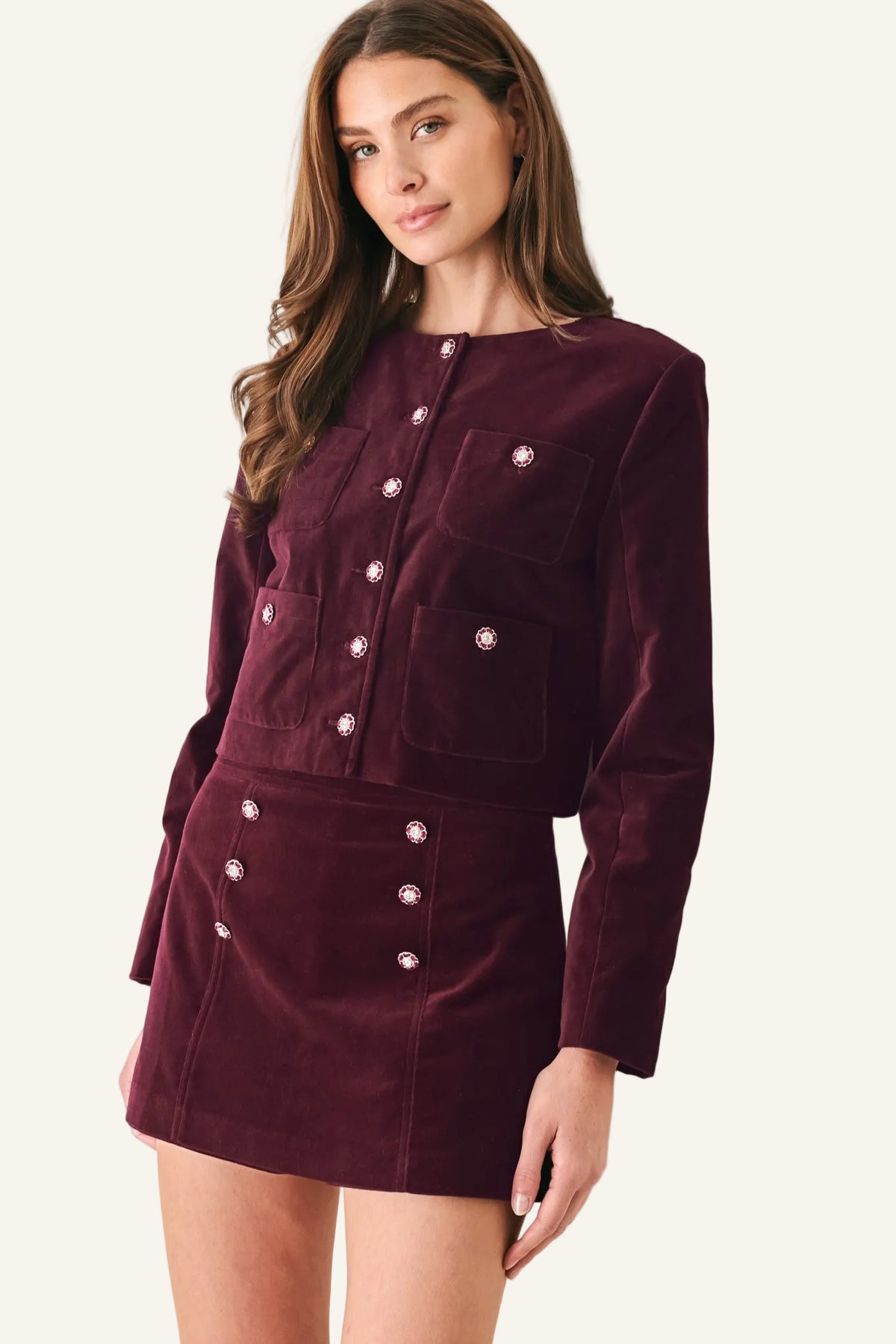A woman wearing a matching burgundy corduroy jacket and mini skirt set with silver-tone decorative buttons, posed against a light gray wall.