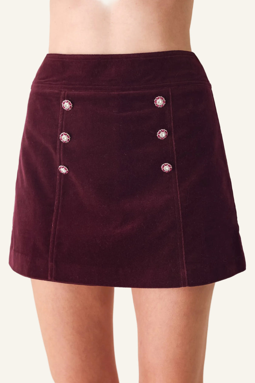A close-up of a model wearing a dark burgundy velvet mini skirt with decorative crystal-embellished buttons on the front.