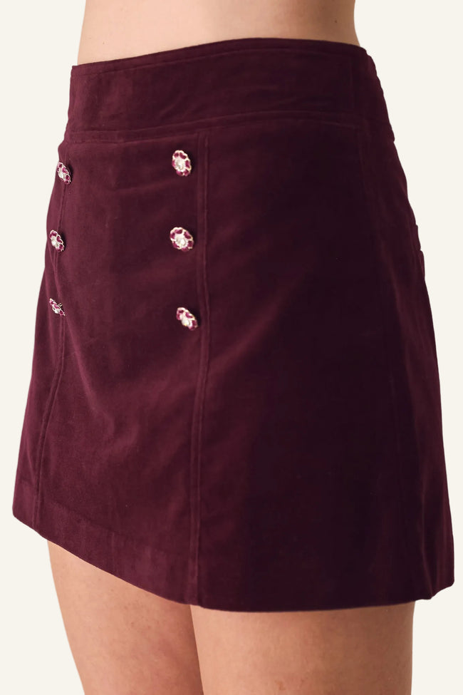 A close-up of a model wearing a dark burgundy velvet mini skirt with decorative pink and white rhinestone buttons on the front.