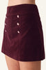 A close-up of a model wearing a dark burgundy velvet mini skirt with decorative pink and white rhinestone buttons on the front.