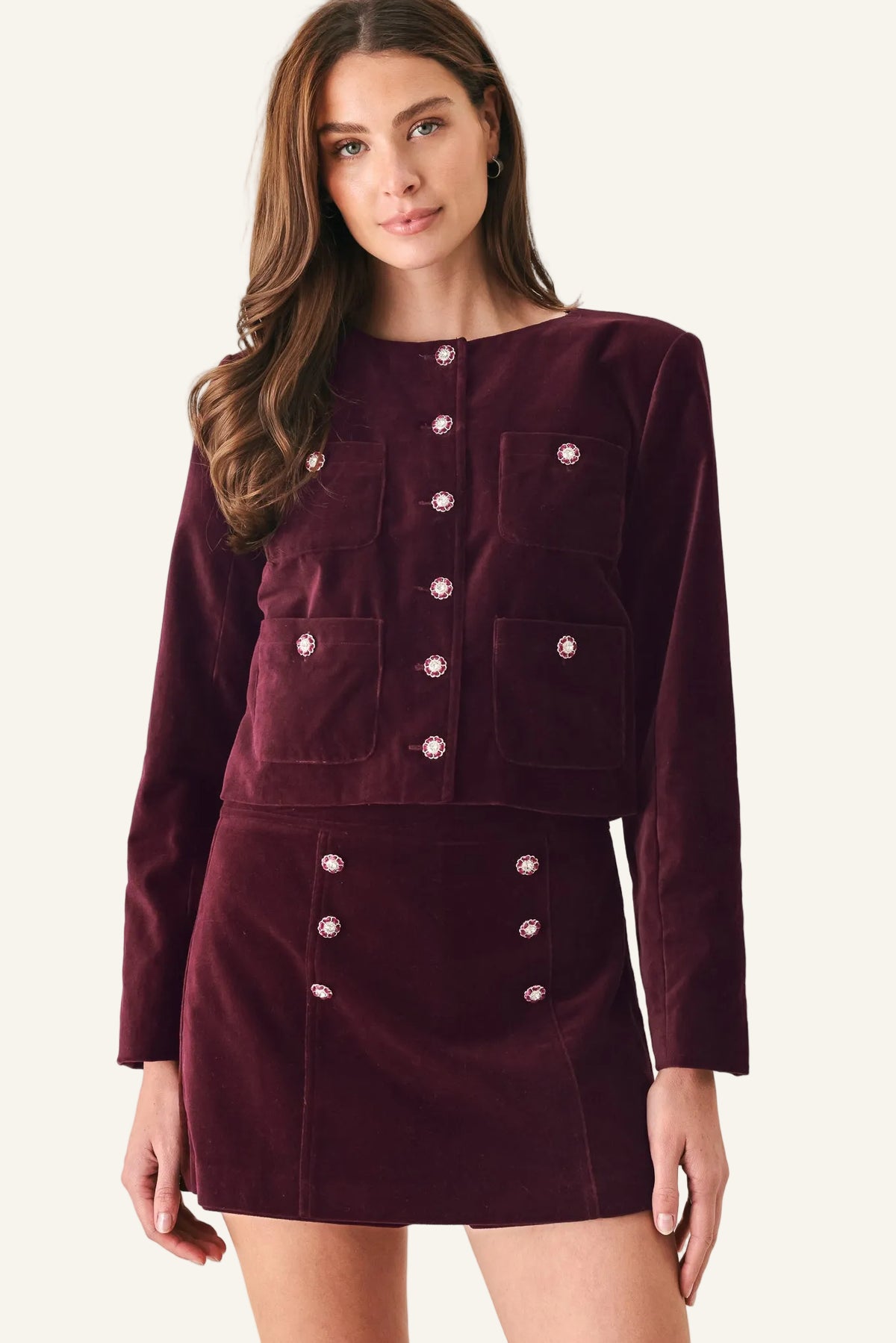 A woman modeling a matching burgundy velvet long-sleeve jacket and mini skirt set with decorative metallic buttons, photographed against a light gray studio background.