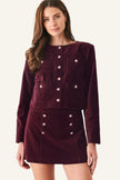 A woman modeling a matching burgundy velvet long-sleeve jacket and mini skirt set with decorative metallic buttons, photographed against a light gray studio background.