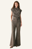 A woman modeling a matching two-piece outfit consisting of a short-sleeve, high-neck, textured taupe top and matching high-waisted wide-leg trousers with a matching belt, set against a plain white studio background.