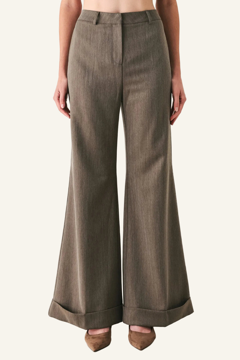A model is wearing a pair of high-waisted, wide-leg trousers in a textured taupe-brown fabric, paired with brown pointed-toe heels against a plain off-white studio background.