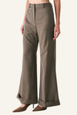 A model is wearing a pair of high-waisted, wide-leg trousers in a textured taupe-brown fabric, paired with brown pointed-toe heels, against a plain light gray studio background.