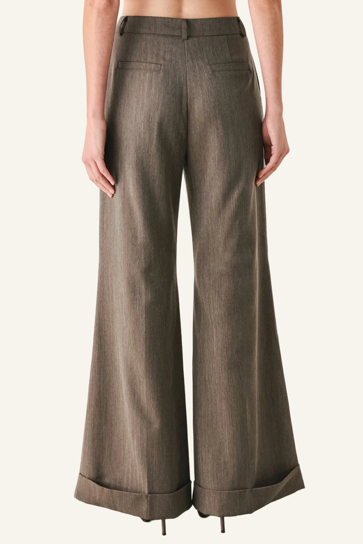 A model shown from the back wearing high-waisted, wide-leg trousers in a textured taupe-brown fabric, styled with black heels against a plain white studio background.