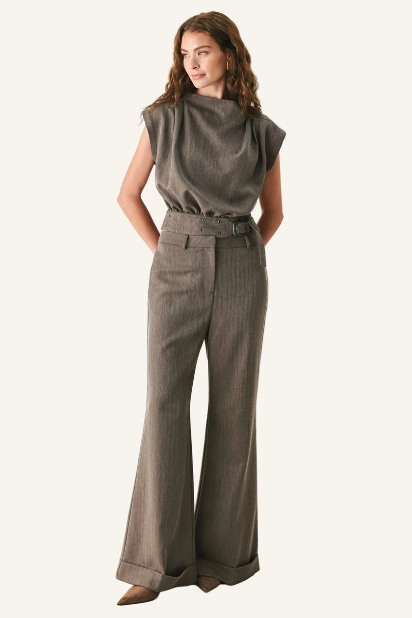 A woman modeling a two-piece outfit consisting of a muted olive-brown sleeveless top and matching high-waisted, wide-leg trousers with a belt, set against a plain light gray studio background.