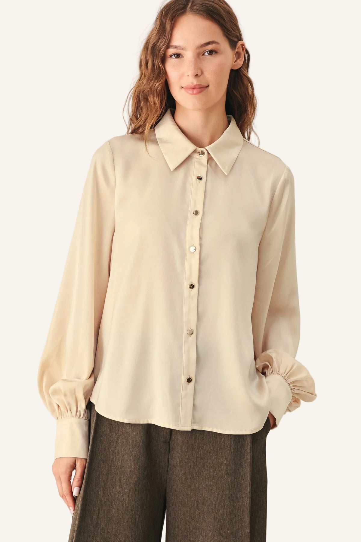 A woman wearing a cream-colored long-sleeve button-up blouse with gathered cuffs, paired with dark brown trousers.