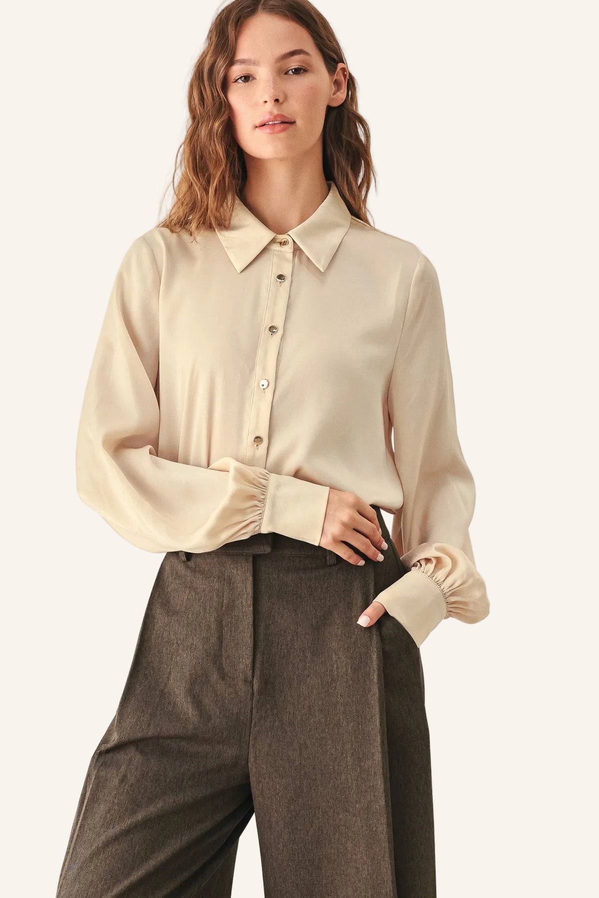 A woman modeling a cream-colored long-sleeve button-up blouse with gathered cuffs, paired with dark brown wide-leg trousers.