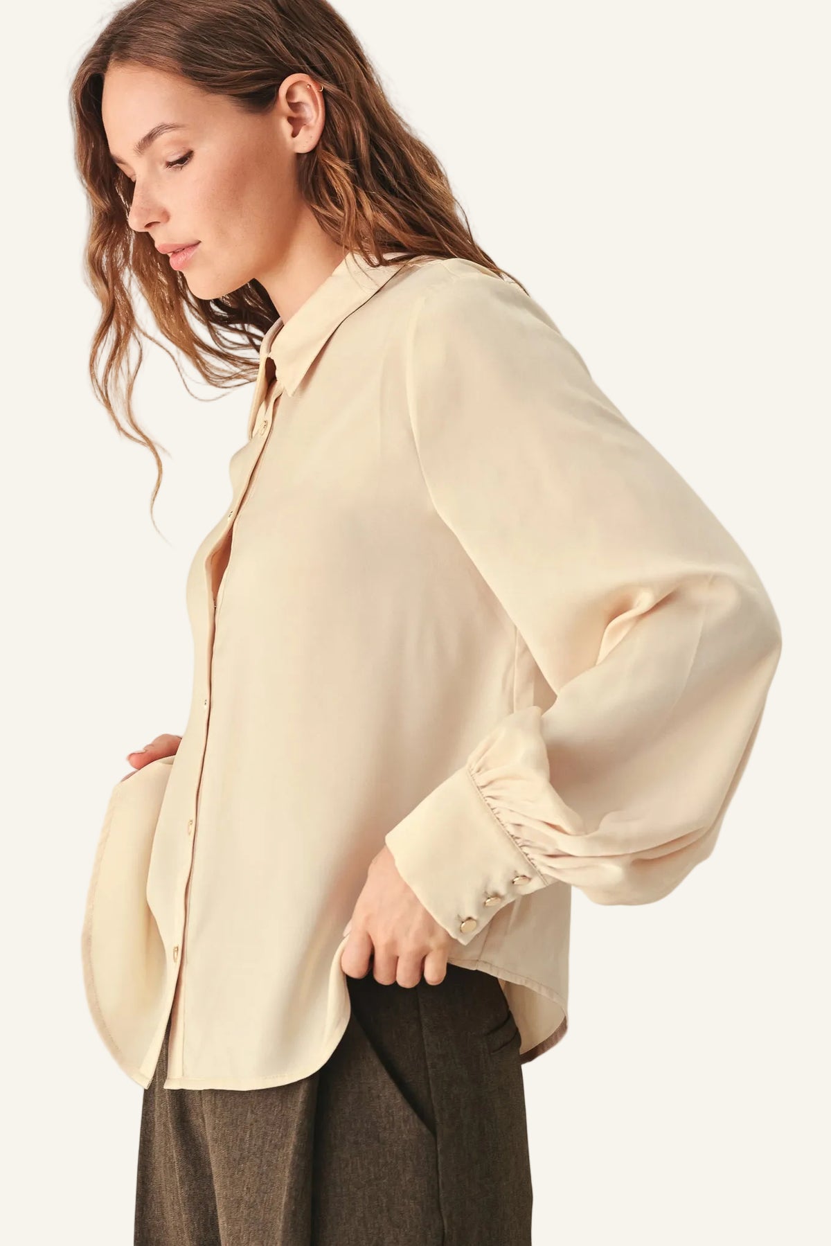A woman wearing a light beige long-sleeve button-up blouse with gathered cuffs, paired with dark brown trousers.