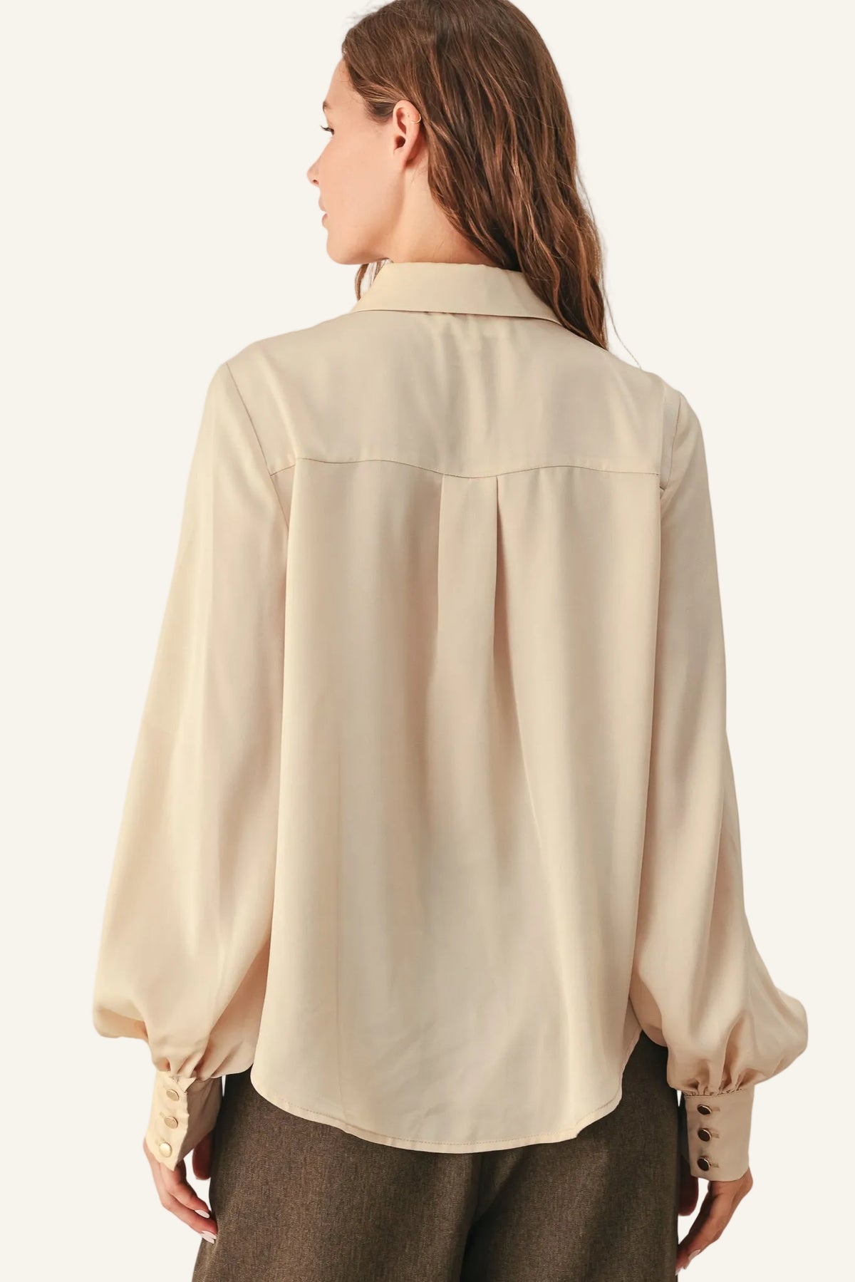 A woman shown from the back wearing a light beige long-sleeve blouse with a subtle sheen and gathered cuffs, paired with brown trousers.