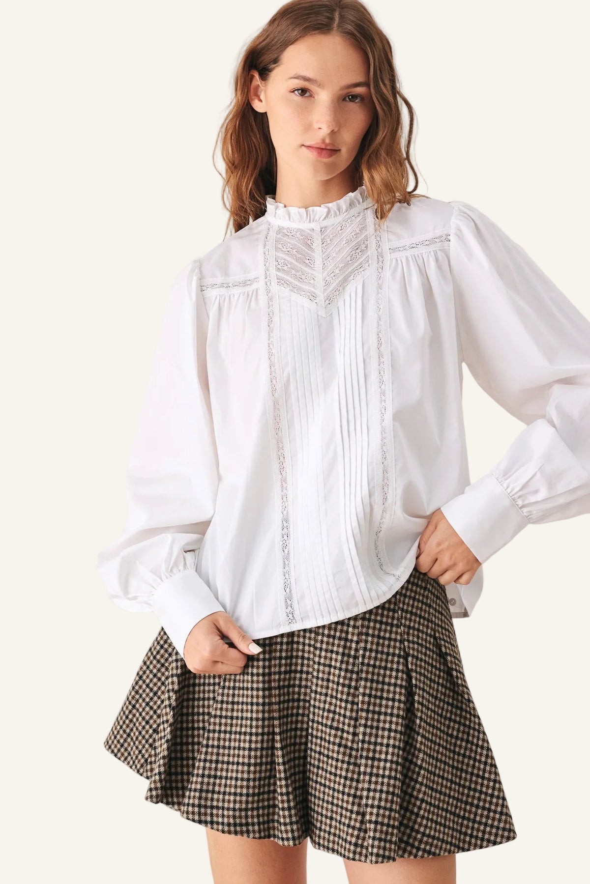 A woman modeling a white long-sleeve blouse with a ruffled high neck, pleated front, and lace detailing, paired with a brown and black gingham pleated mini skirt.