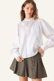 A woman modeling a white long-sleeve blouse with a ruffled high neck, pleated front, and lace detailing, paired with a brown and black gingham pleated mini skirt.