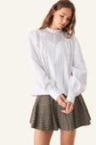 A woman wearing a white long-sleeve blouse with delicate embroidery and a ruffled high neck, paired with a brown and black checkered pleated skirt.