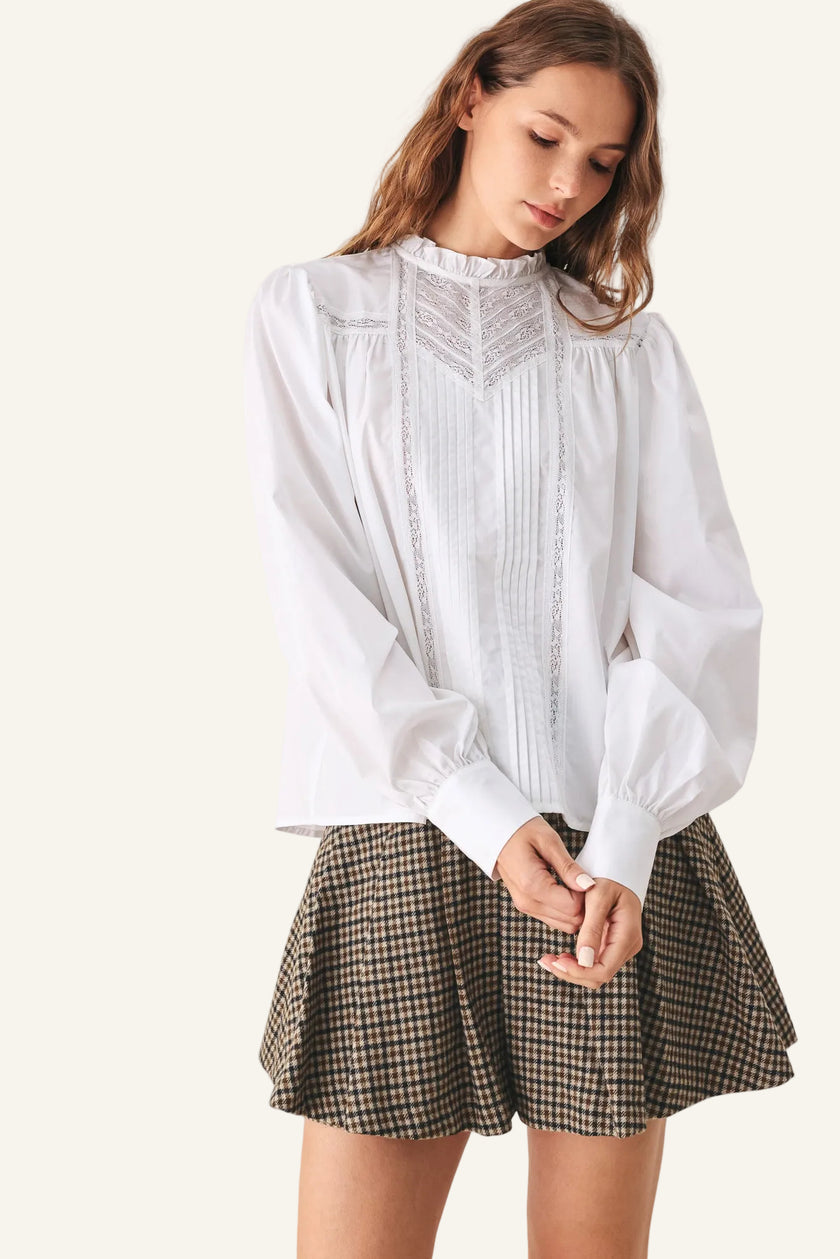A woman wearing a white long-sleeve blouse with delicate embroidery and a ruffled high neck, paired with a brown and black checkered pleated skirt.