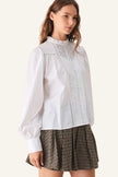 A woman wearing a white long-sleeve blouse with a ruffled high neck and vertical lace-like front detailing, paired with a brown and beige checkered pleated mini skirt.