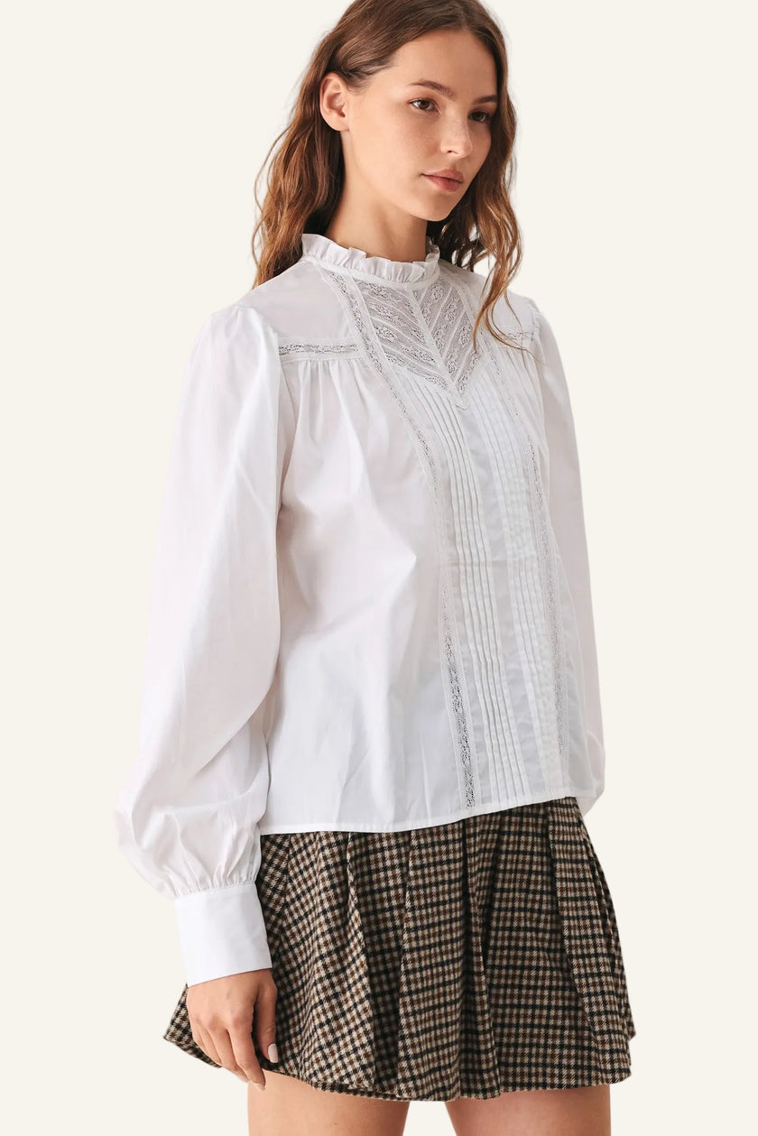 A woman wearing a white long-sleeve blouse with a ruffled high neck and vertical lace-like front detailing, paired with a brown and beige checkered pleated mini skirt.