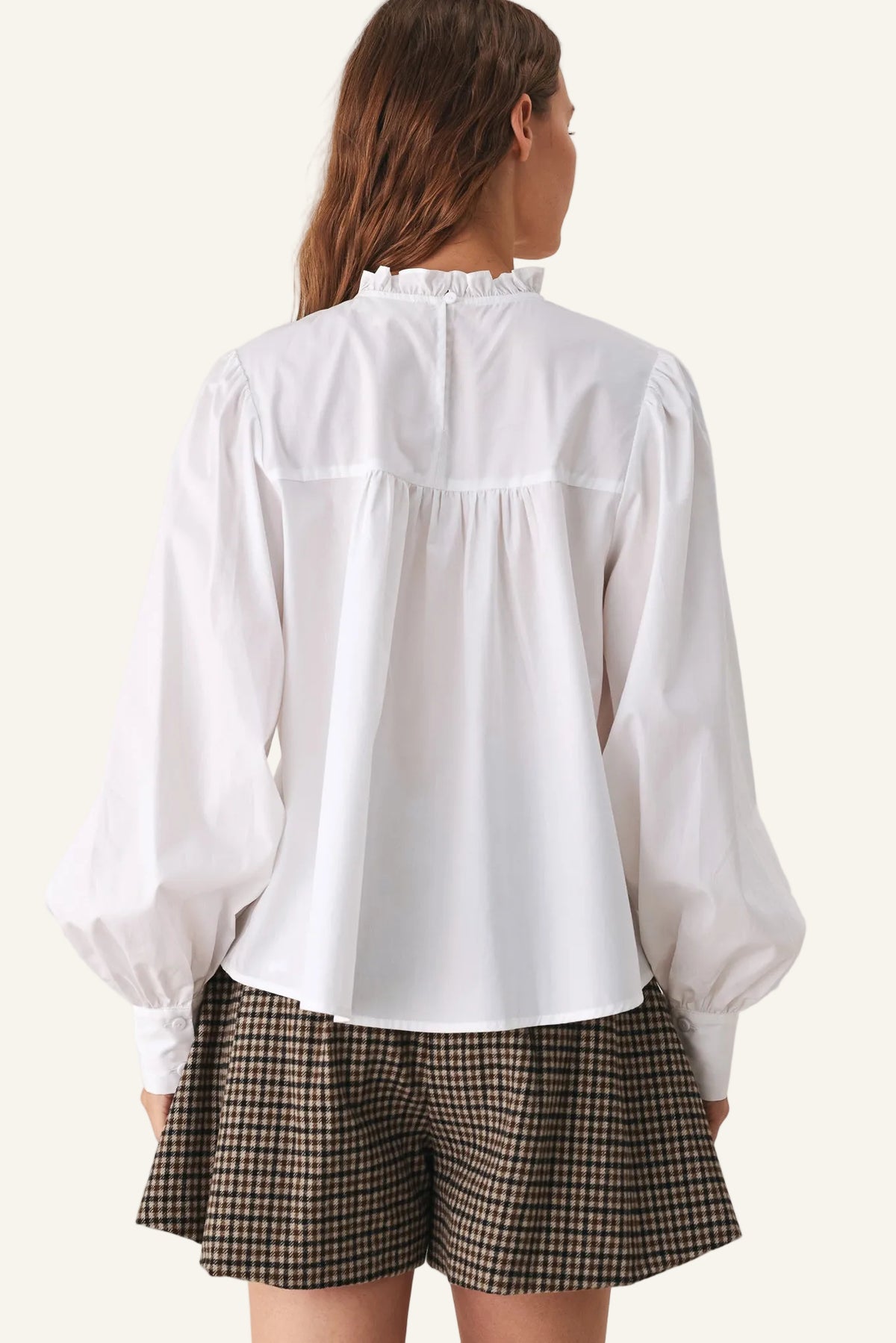 A woman shown from the back wearing a white long-sleeve blouse with a ruffled high neck and gathered waist, paired with brown and black checkered shorts.