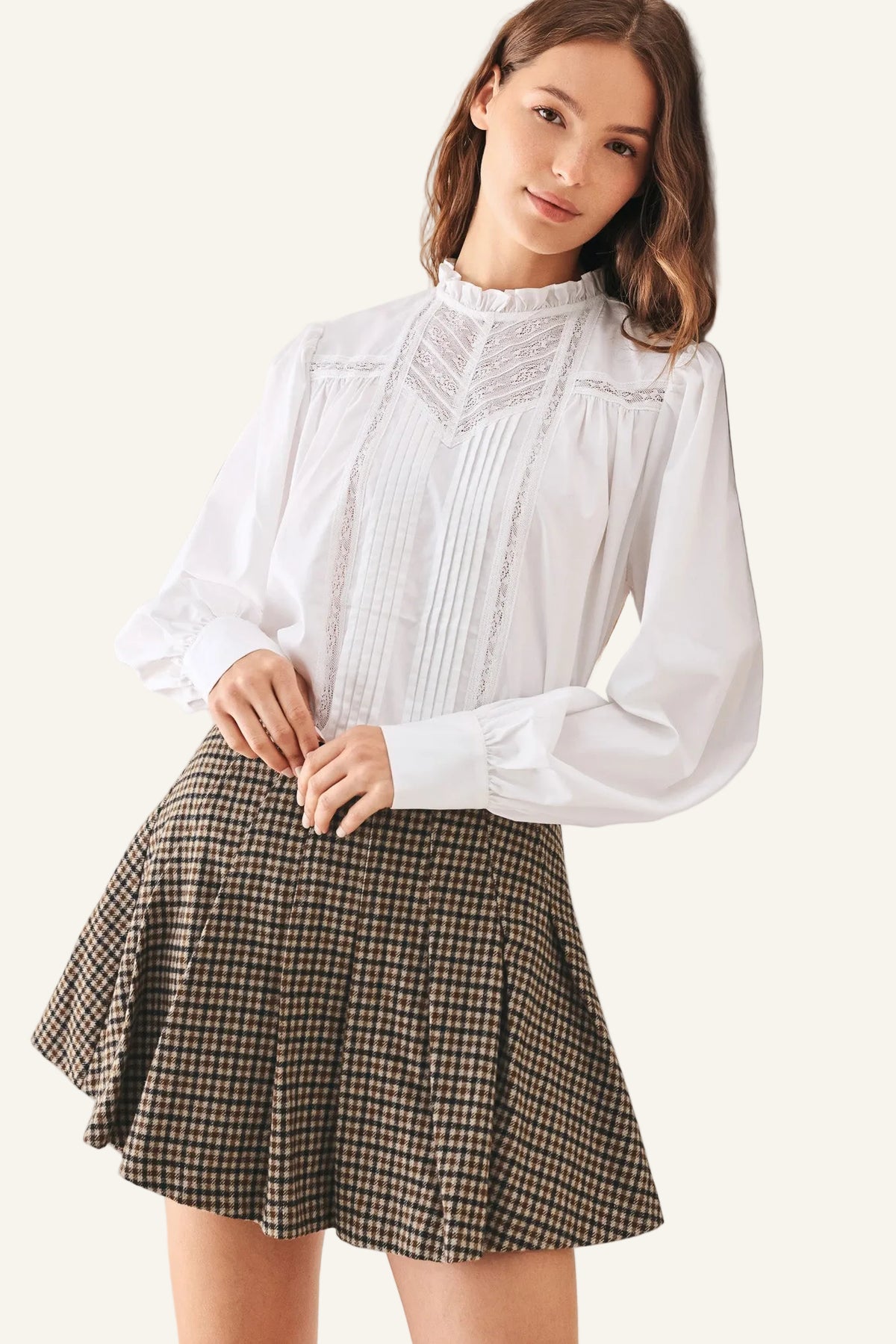 A woman wearing a white long-sleeve blouse with pleated detailing and a brown and beige houndstooth mini skirt.