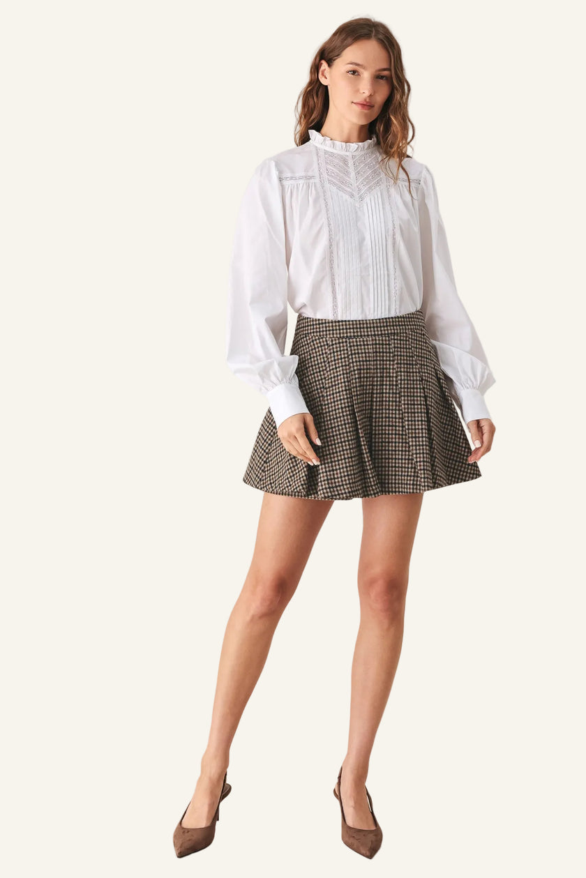 A woman modeling a white long-sleeve blouse with a ruffled high neck and pleated front, paired with a brown and beige houndstooth mini skirt and brown pointed-toe heels.