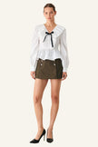 A woman modeling a white long-sleeve blouse with a ruffled collar and black bow tie detail, paired with an olive green mini skirt and black heels, against a plain light gray studio background.