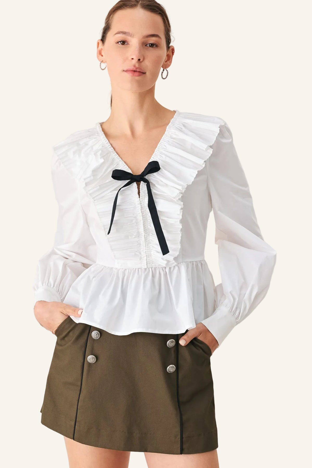 A woman modeling a white long-sleeve blouse with a ruffled V-neck, gathered waist, and a black ribbon bow at the center, paired with an olive green mini skirt against a light beige studio background.