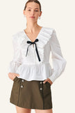 A woman modeling a white long-sleeve blouse with a ruffled V-neck, gathered waist, and a black ribbon bow at the center, paired with an olive green mini skirt against a light beige studio background.