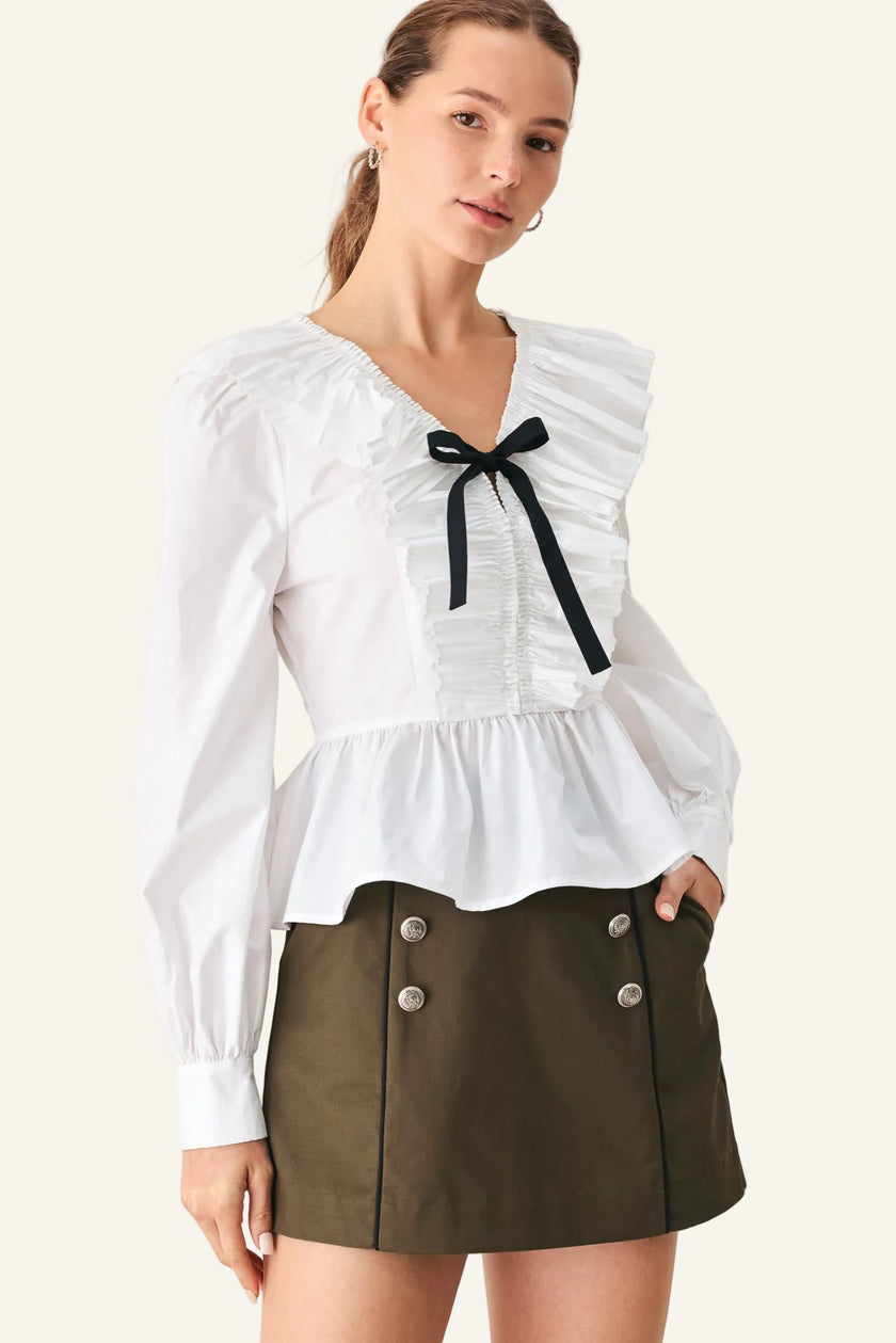 A woman modeling a white long-sleeve blouse with ruffled shoulders, a smocked front, and a black ribbon bow at the neckline, paired with an olive green mini skirt against a light beige studio background.