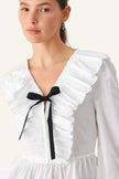 A close-up of a model wearing a white long-sleeve blouse with dramatic ruffles, a deep V-neckline, and a black ribbon bow tied at the center chest.