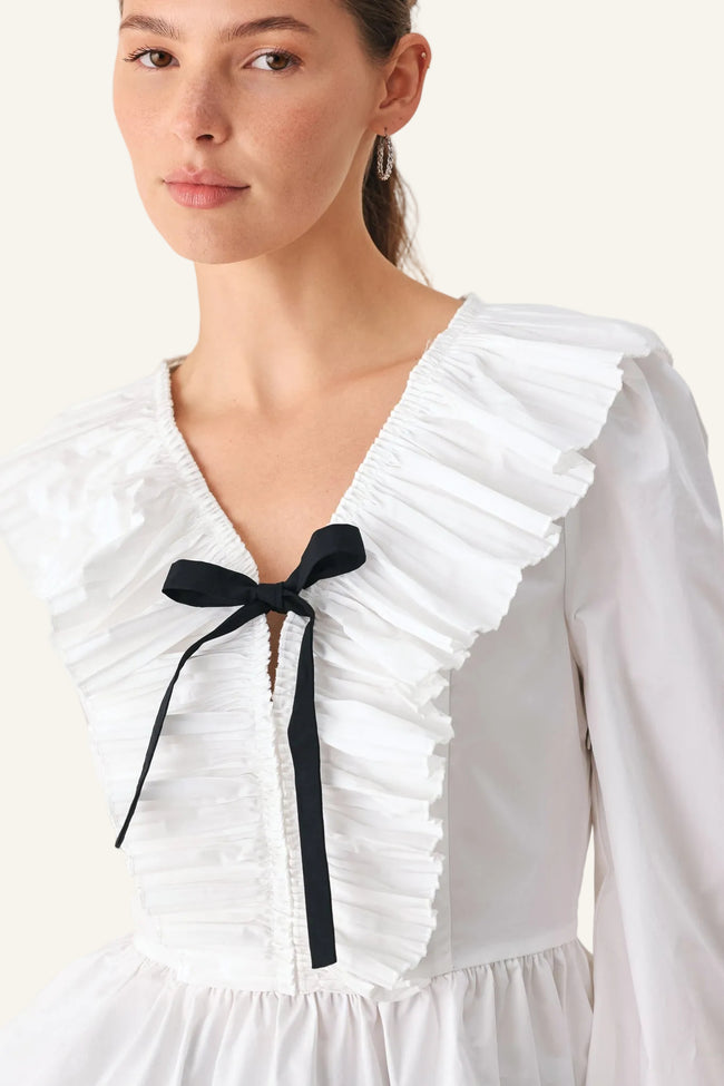 A close-up of a model wearing a white long-sleeve blouse with dramatic ruffles, a deep V-neckline, and a black ribbon bow tied at the center chest.