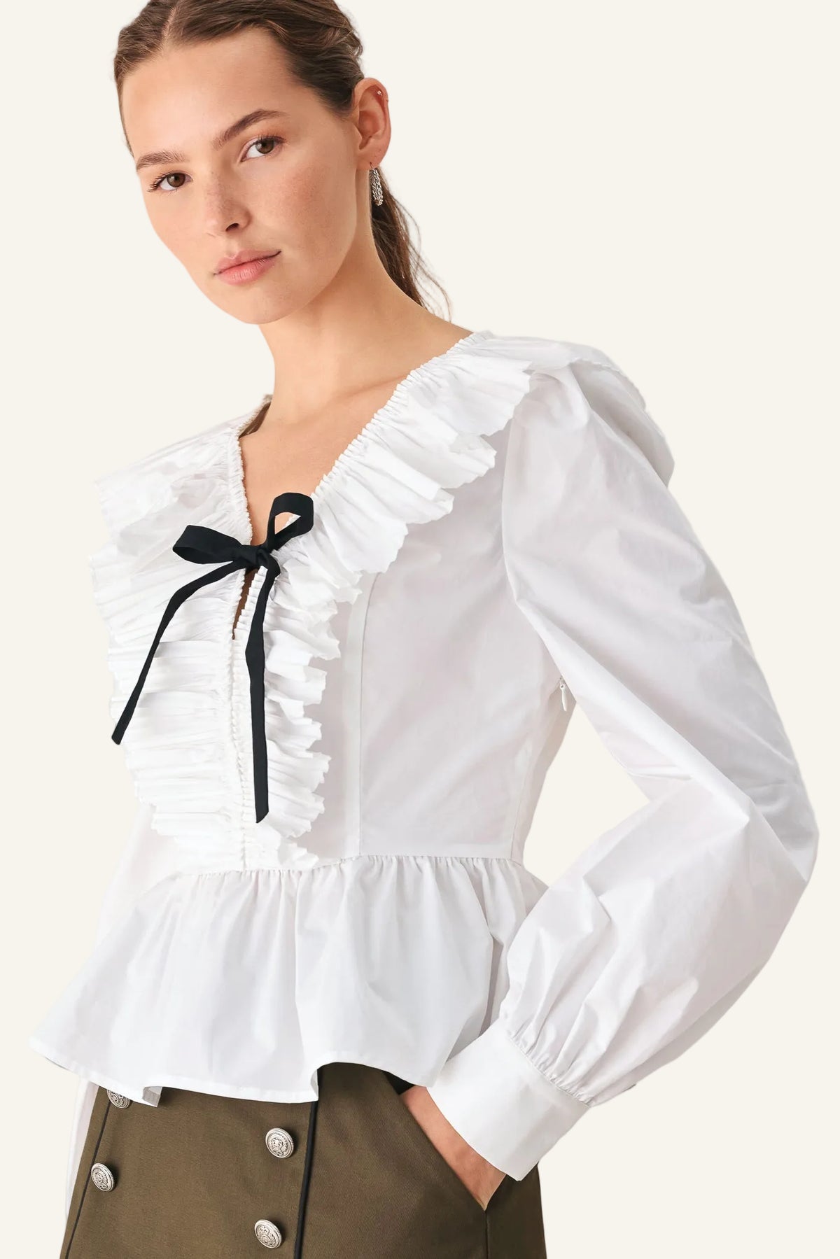 A woman wearing a white long-sleeve blouse with ruffled trim, a black ribbon bow at the neckline, and a gathered peplum hem, paired with dark olive pants against a light studio background.
