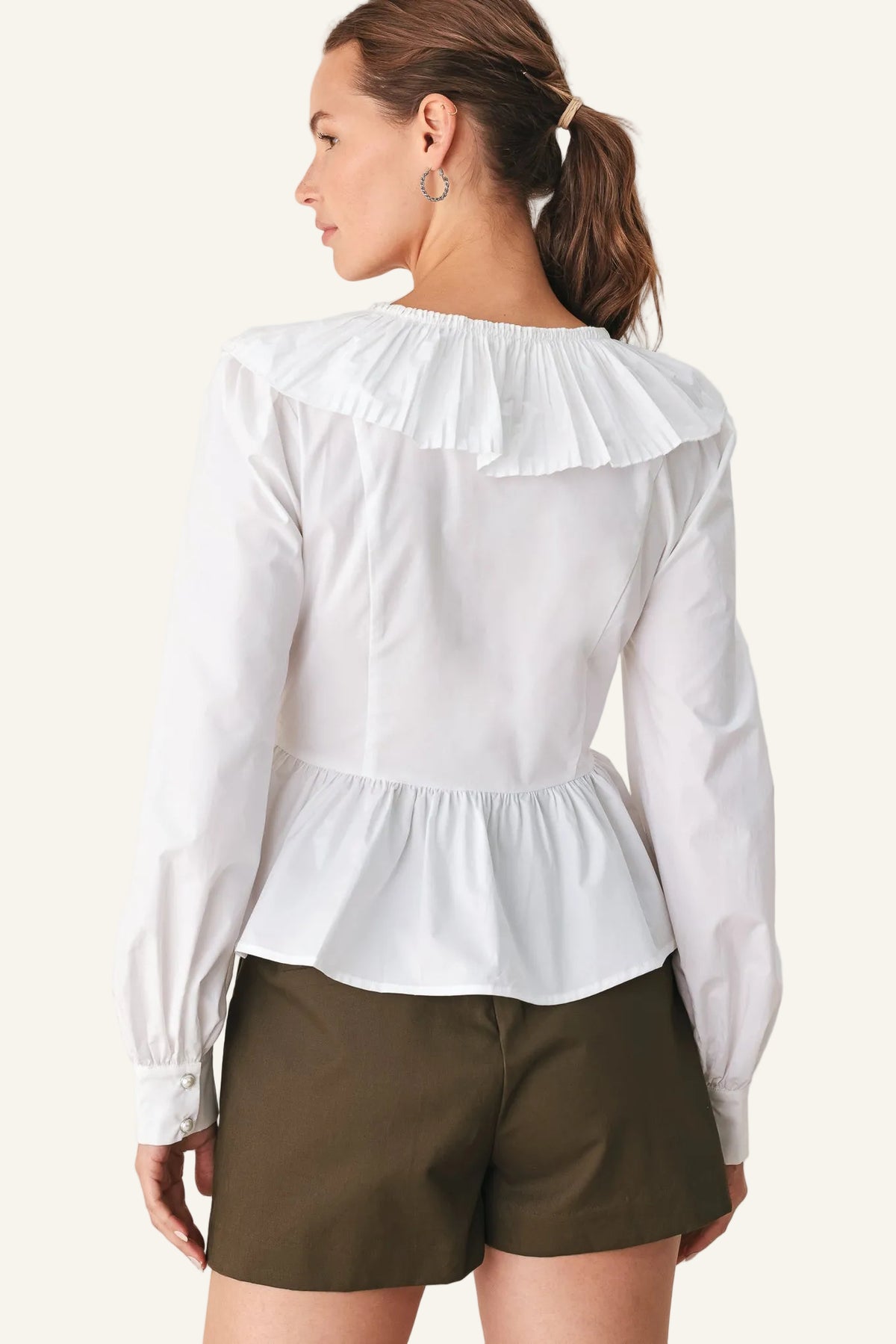 A woman shown from the back wearing a white long-sleeve blouse with a ruffled collar and peplum waist, paired with olive green shorts, against a plain light background.
