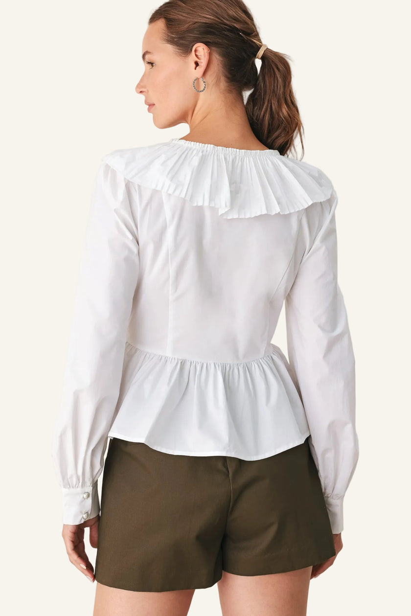 A woman shown from the back wearing a white long-sleeve blouse with a ruffled collar and peplum waist, paired with olive green shorts, against a plain light background.