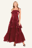 A woman modeling a sleeveless, tiered maxi dress in a shiny deep burgundy or maroon satin-like fabric, paired with black heeled sandals.