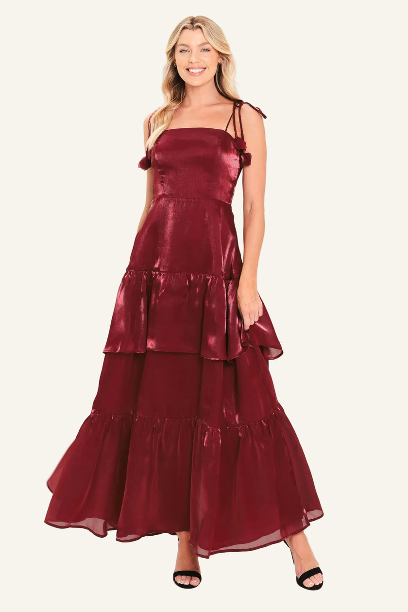 A woman modeling a sleeveless, tiered maxi dress in a shiny deep burgundy or maroon satin-like fabric, paired with black heeled sandals.