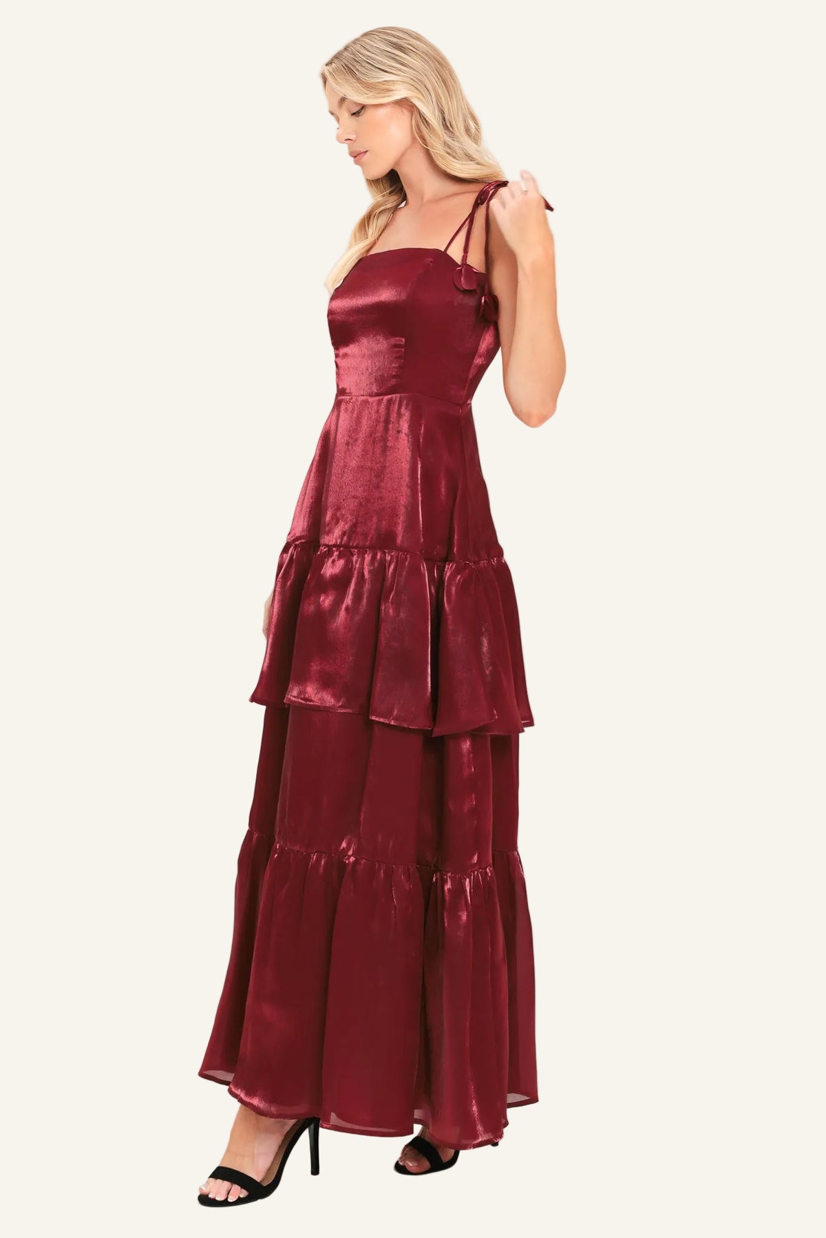 A woman modeling a sleeveless, tiered maxi dress in a deep burgundy satin fabric, paired with black heeled sandals.