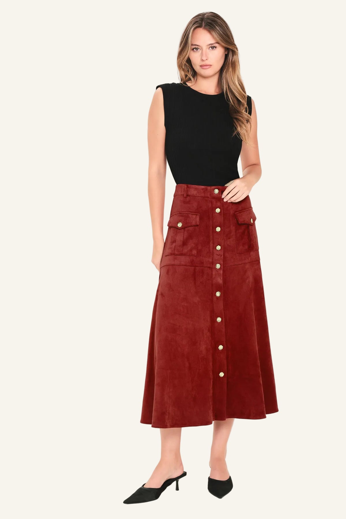 A woman modeling a high-waisted, mid-length rust red corduroy skirt with gold-tone button details down the front, paired with a black sleeveless top and black pointed-toe mule heels, against a plain off-white studio background.