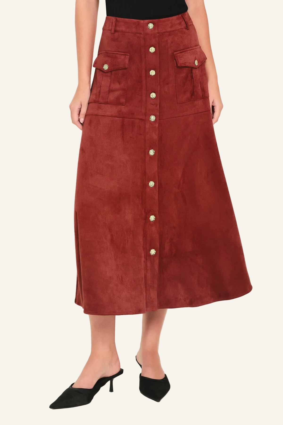 A rust-brown suede midi skirt with a button-down front, flap pockets, and gold-tone button accents, shown on a model wearing black pointed-toe mule heels against a light studio background.