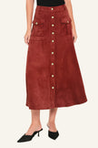 A rust-brown suede midi skirt with a button-down front, flap pockets, and gold-tone button accents, shown on a model wearing black pointed-toe mule heels against a light studio background.