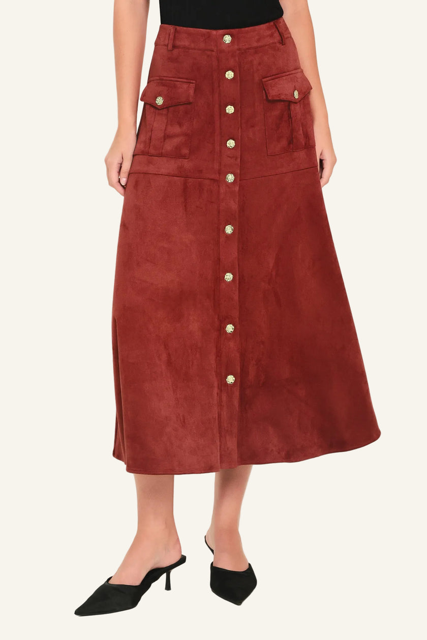 A rust-brown suede midi skirt with a button-down front, flap pockets, and gold-tone button accents, shown on a model wearing black pointed-toe mule heels against a light studio background.