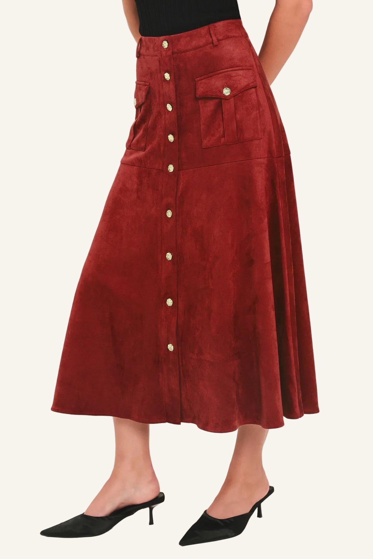 A model wearing a high-waisted, rust-red corduroy midi skirt with a button-down front and flap pockets, paired with black pointed-toe mule heels against a plain white studio background.