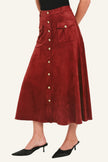 A model wearing a high-waisted, rust-red corduroy midi skirt with a button-down front and flap pockets, paired with black pointed-toe mule heels against a plain white studio background.