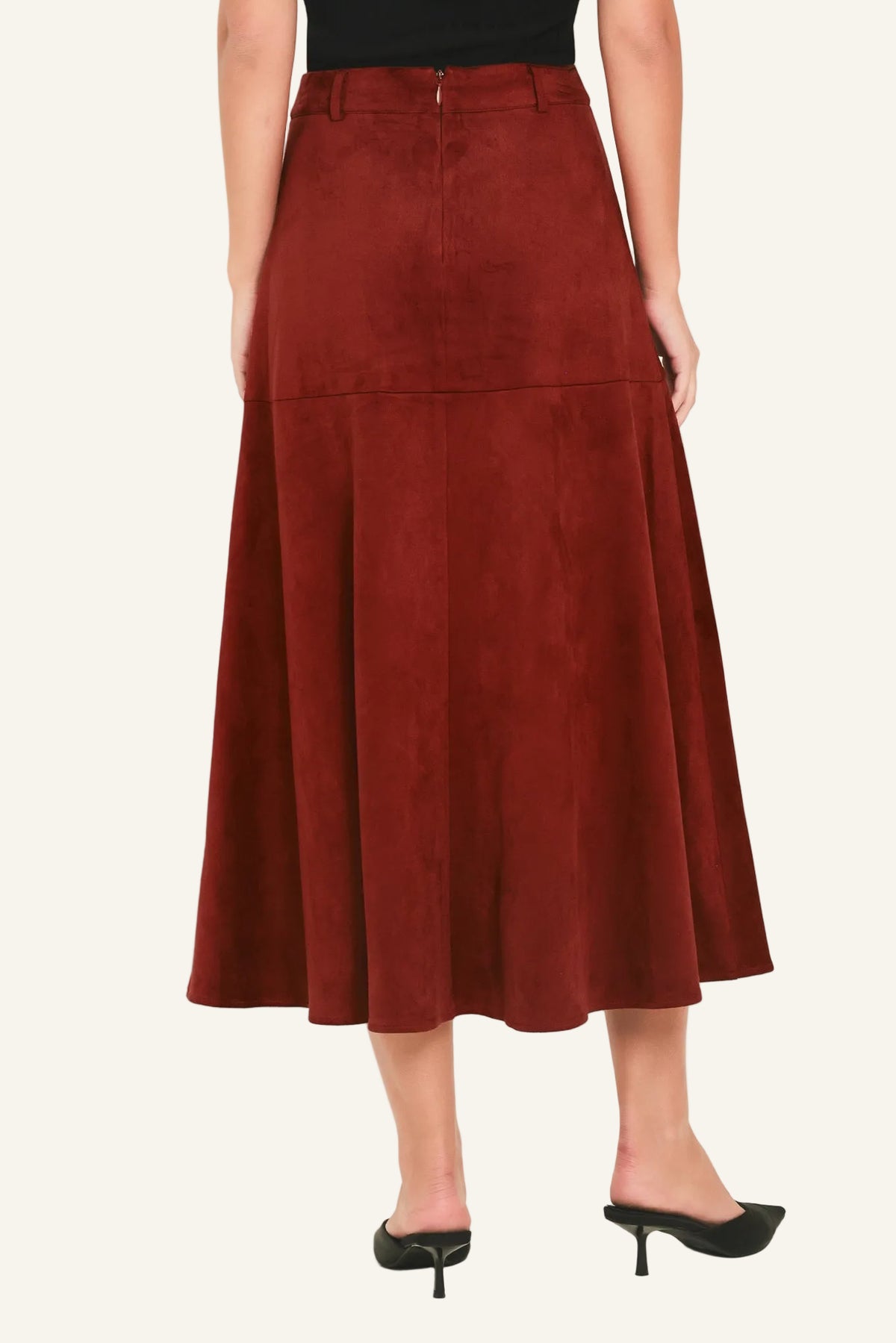 A woman wearing a rust-brown suede midi skirt with a black top and black pointed-toe mule heels, shown from the waist down against a plain off-white studio background.